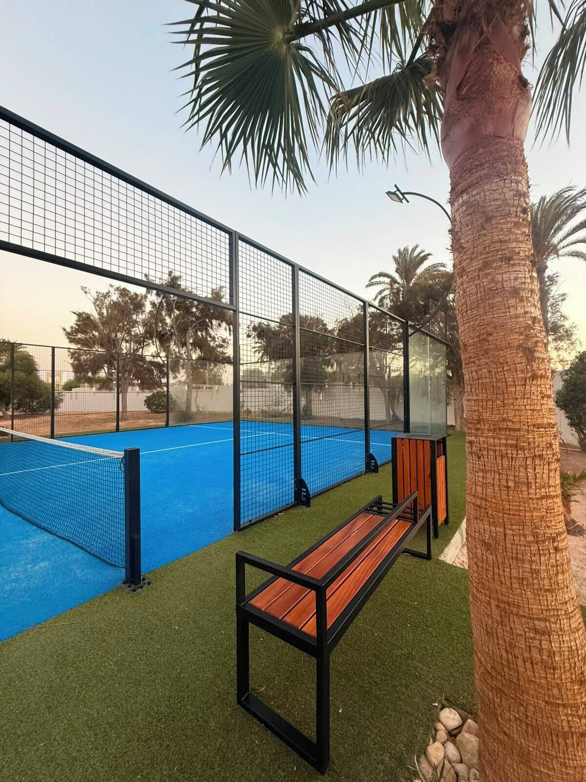 A tennis court with a net, surrounded by a metal fence, and a bench in the foreground.