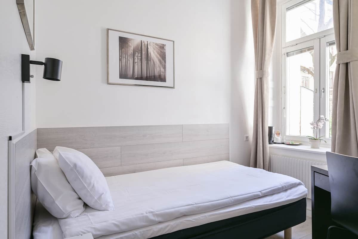 A neatly made bed with white linens, a light wood headboard, a framed picture on the wall, and a window with curtains.