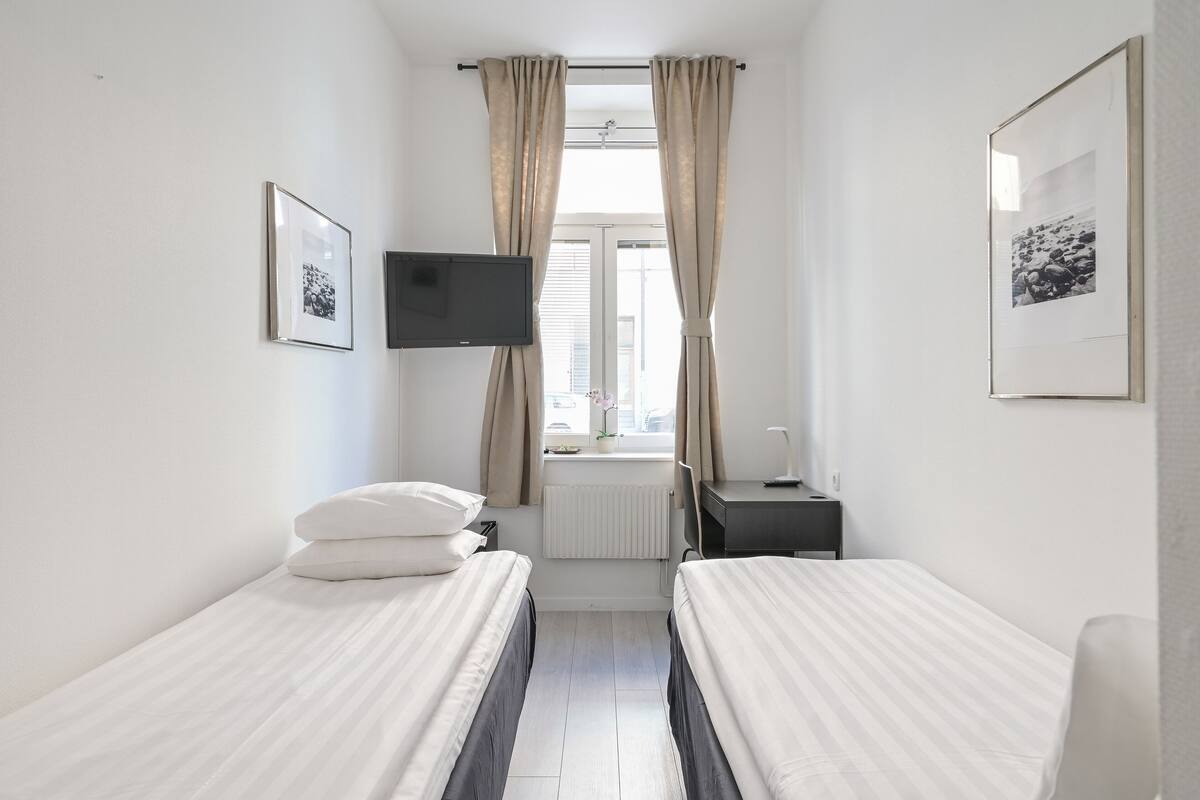 A small, clean room with two single beds, a flat-screen TV mounted on the wall, a window with curtains, and a radiator.