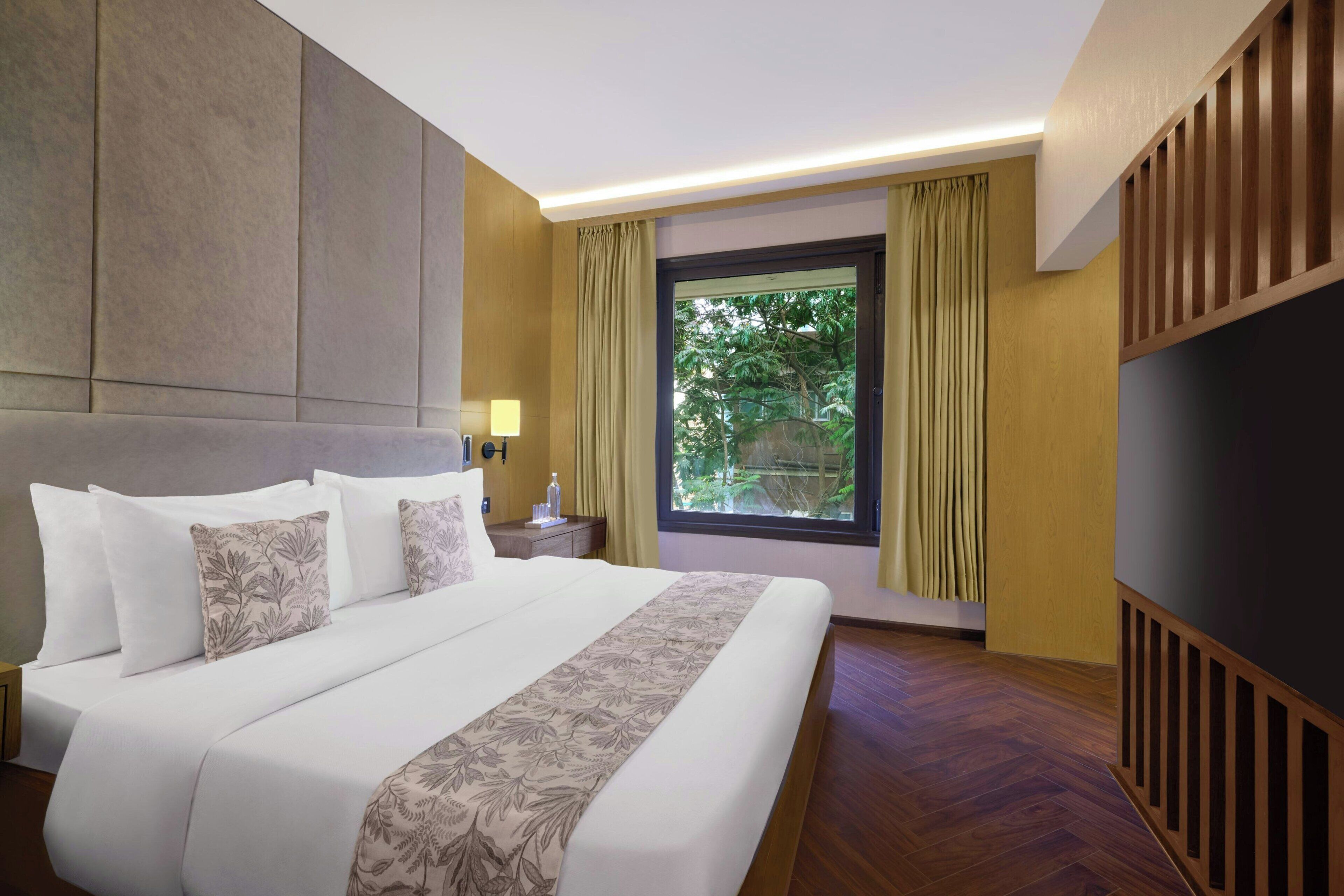 Habitación de hotel con una cama grande, una mesita de noche, una ventana con cortinas y vistas a zonas verdes.