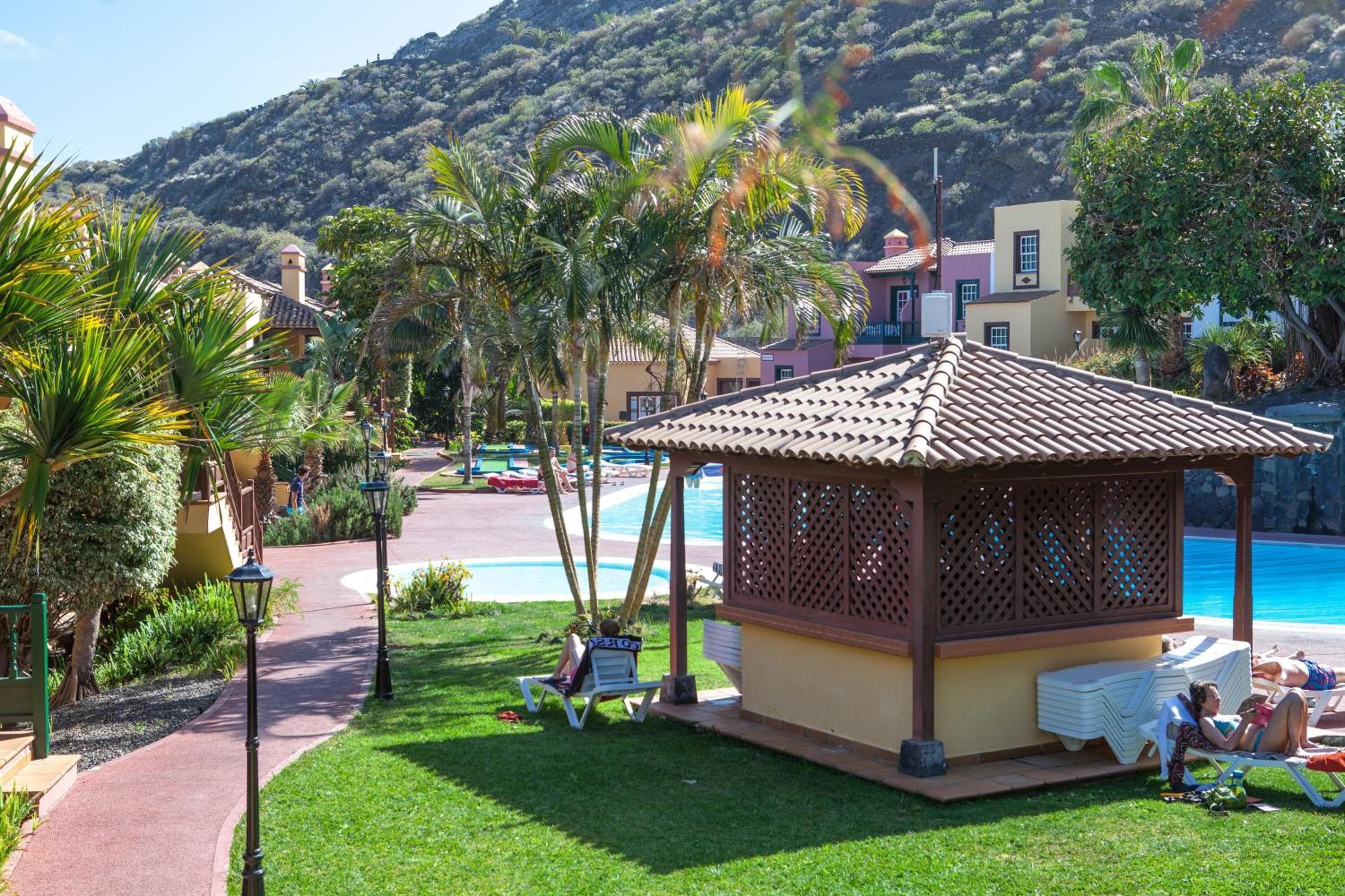 Un pabellón con techo de tejas, rodeado por una piscina y palmeras, con una montaña al fondo.