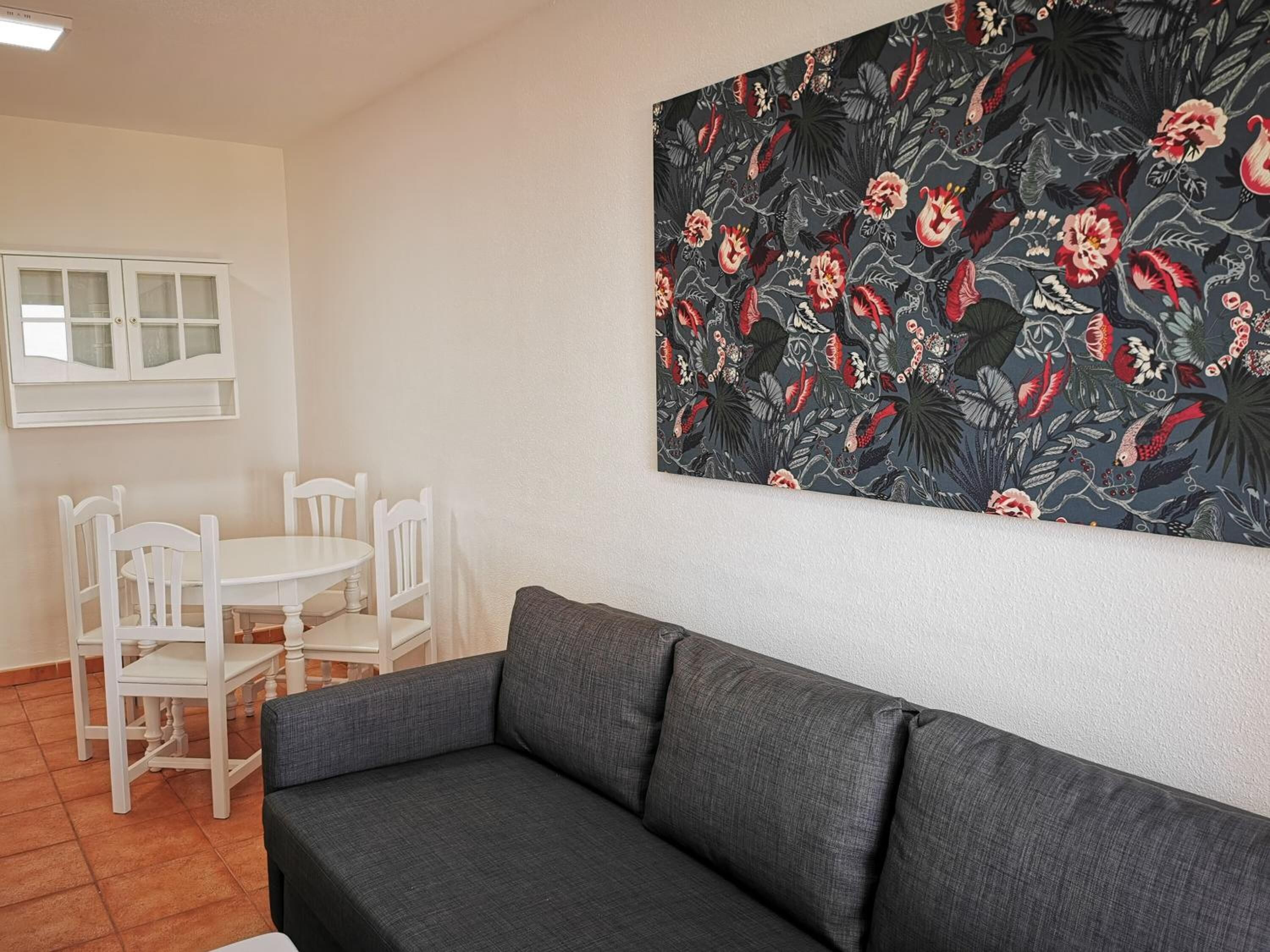 A living room with a grey sofa, a dining table with white chairs, and a wall with a floral pattern.