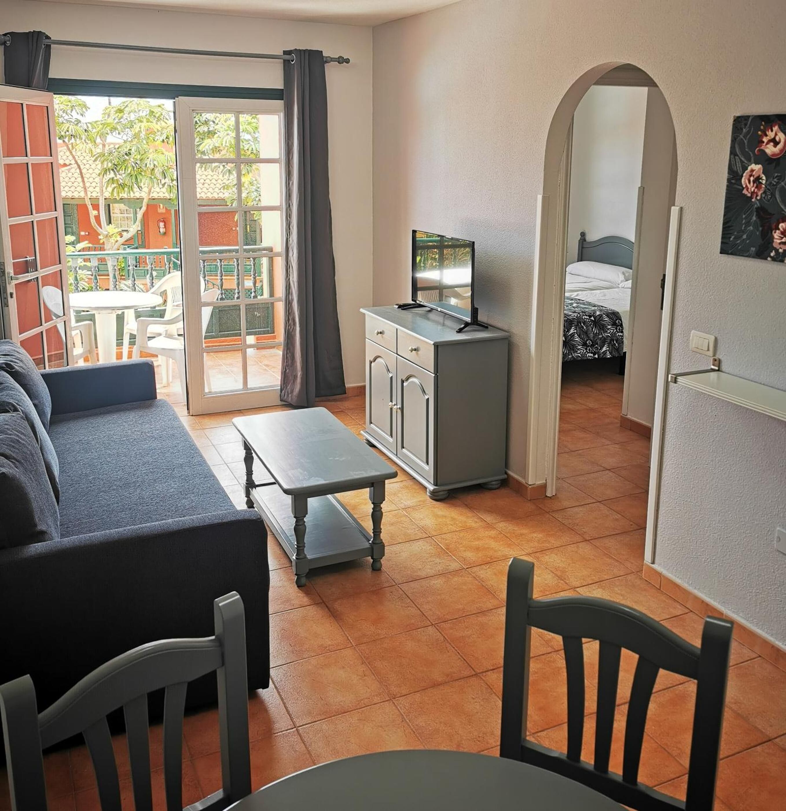 Un salon confortable avec un canapé, une petite table et un téléviseur posé sur un meuble. Il y a un balcon avec une table et des chaises à l'extérieur.
