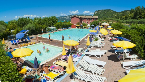 Ein Poolbereich mit mehreren Liegestühlen und Sonnenschirmen, umgeben von Grünflächen und einem Gebäude im Hintergrund.