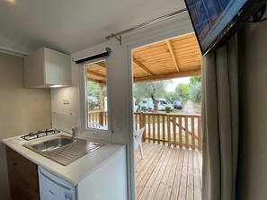 A compact kitchen with a sink, a stove, and a small refrigerator. There is a wooden deck with a view of parked cars and greenery outside.