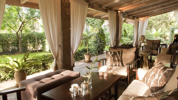 A covered outdoor seating area with a wooden table, cushioned chairs, and a view of a lush garden. - Palacina The Residence & The Suites (Nairobi)