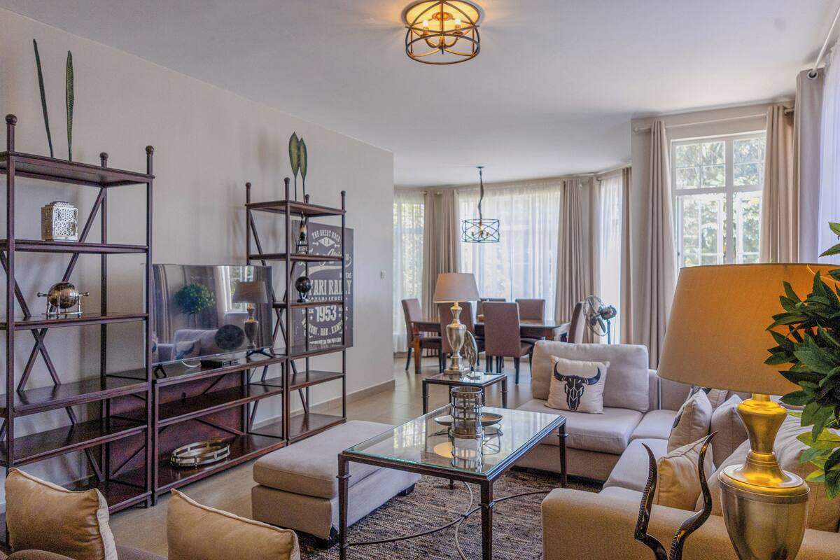 A living room with a sofa, armchairs, a coffee table, and a bookshelf. There is a dining area with chairs and a table in the background.