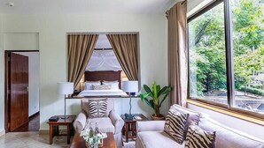 A hotel room with a bed, two sofas, a coffee table with fruit, a potted plant, and a window with a view of greenery. - Palacina The Residence & The Suites (Nairobi)