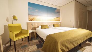 Una habitación de hotel con cama, una silla, una mesita de noche, una lámpara y un cuadro grande con un paisaje en la pared.