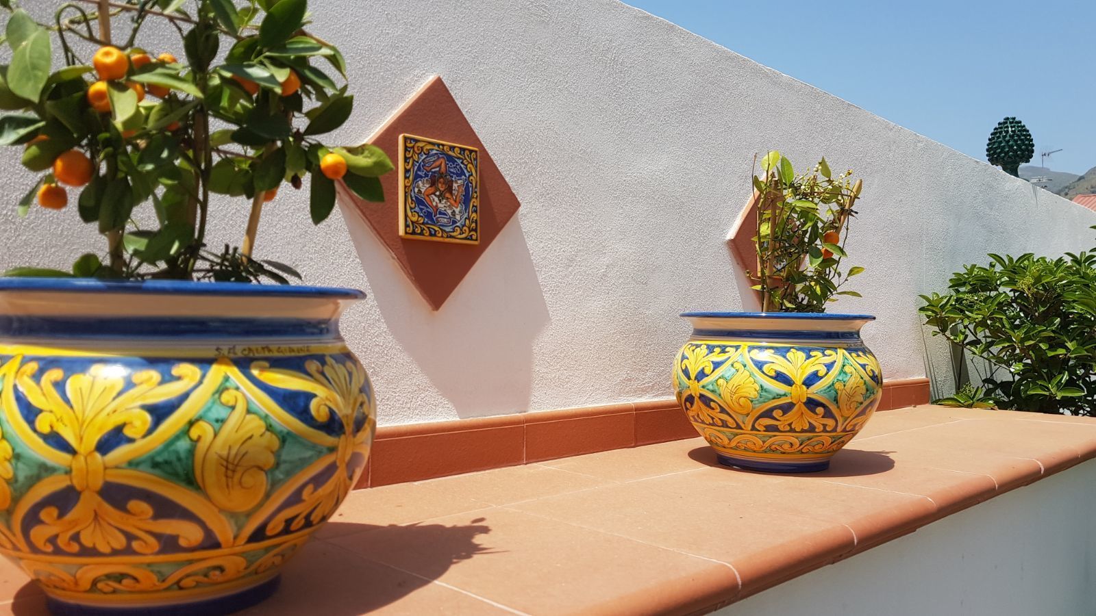Dos macetas grandes y ornamentadas de cerámica sobre un repisa de baldosas frente a una pared blanca con un azulejo decorativo.