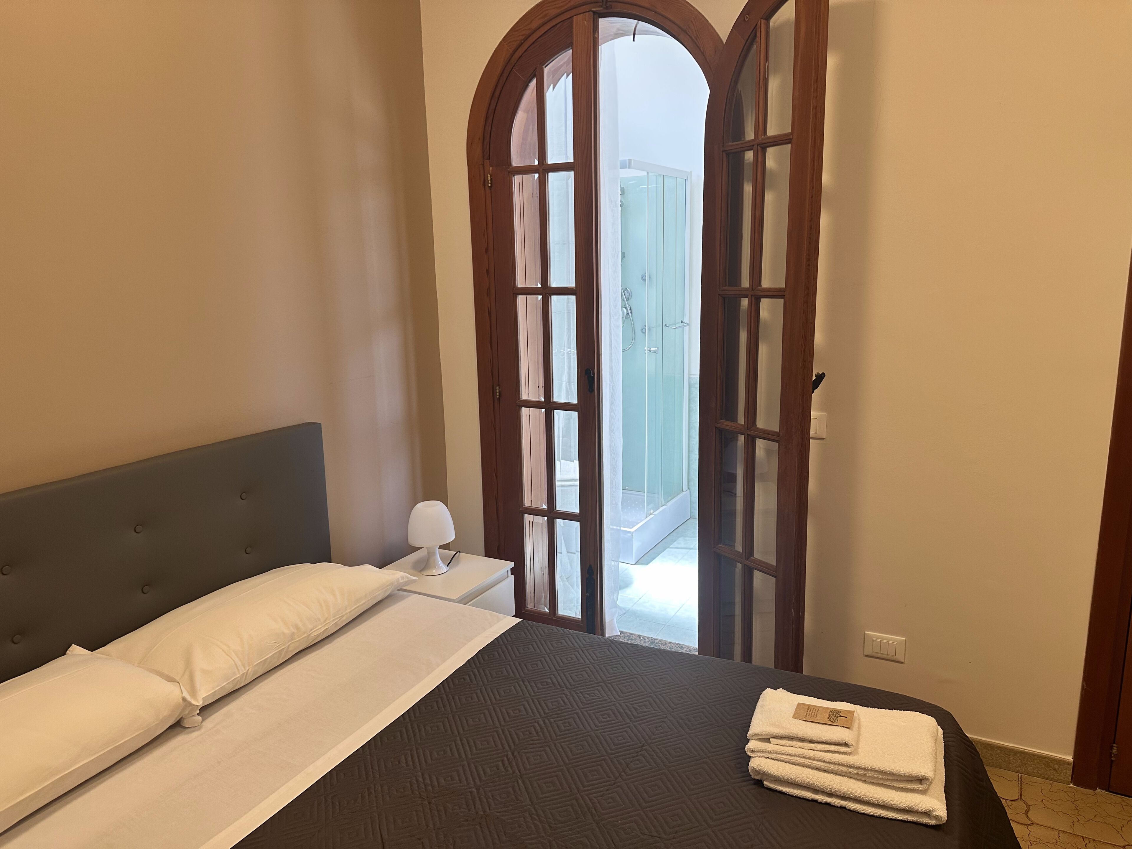 A bedroom with a bed, a nightstand, a lamp, and a view of a balcony through an arched door.