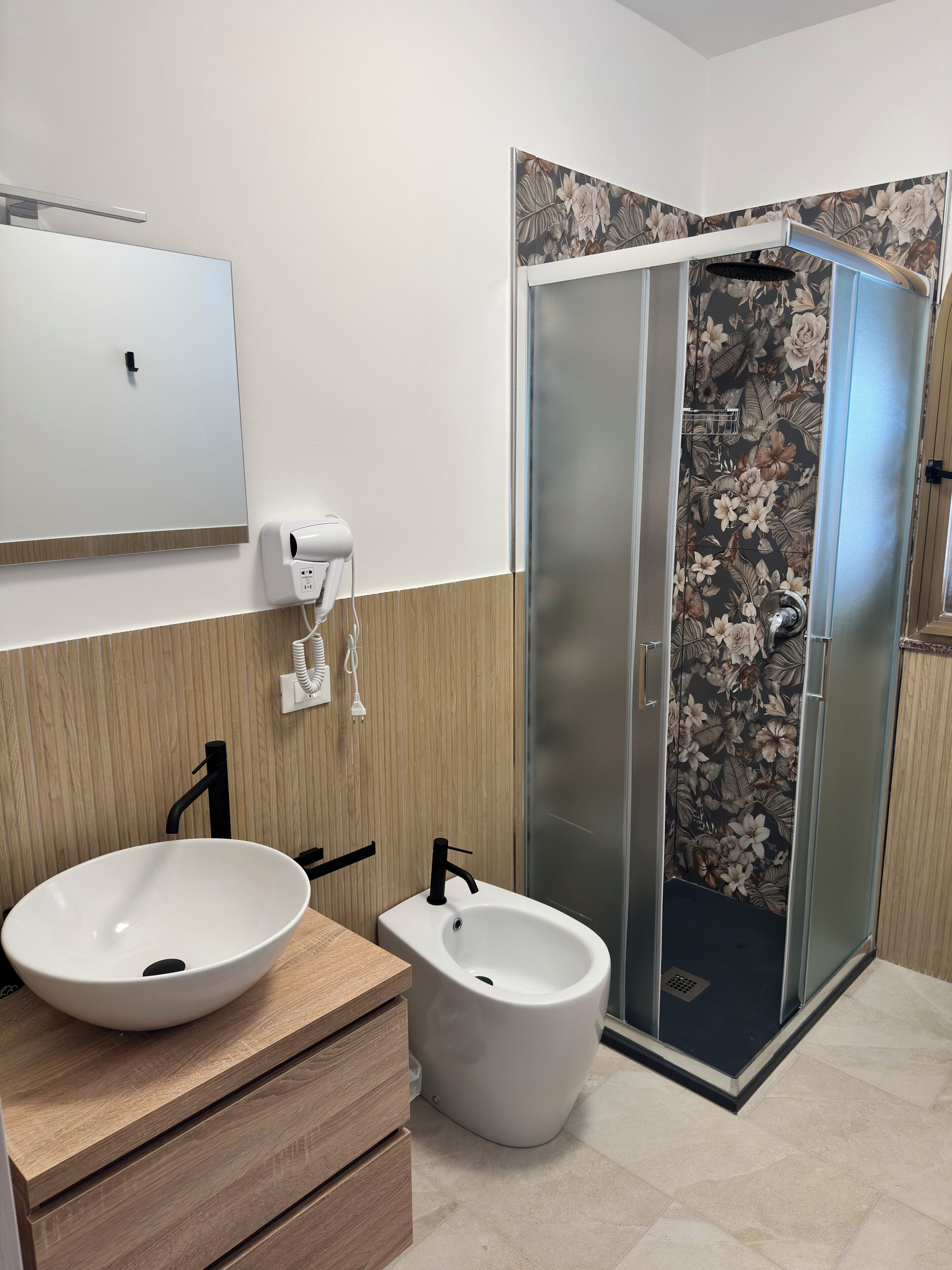 A modern bathroom with a large white sink, a bidet, a mirror, and a shower with floral wallpaper.
