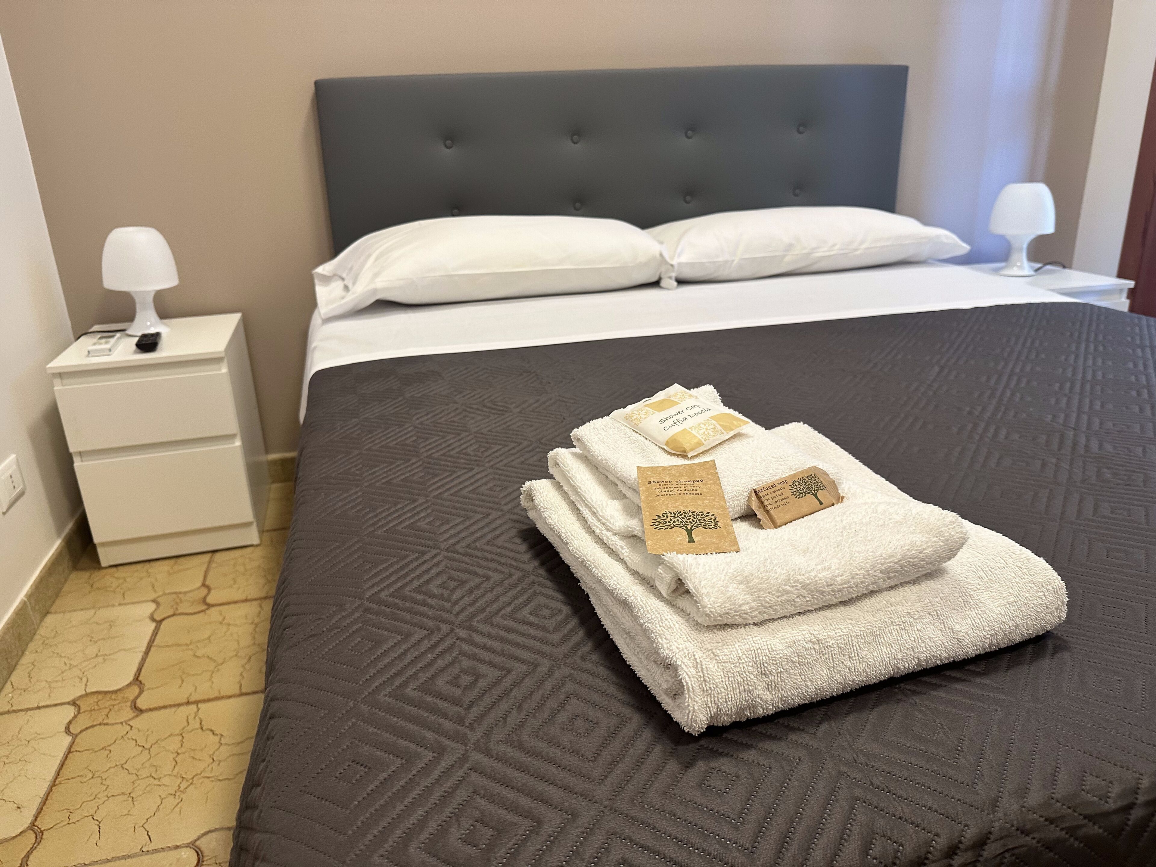 A hotel room with a neatly made bed, a bedside table with a lamp, and a folded towel with a tag on it.