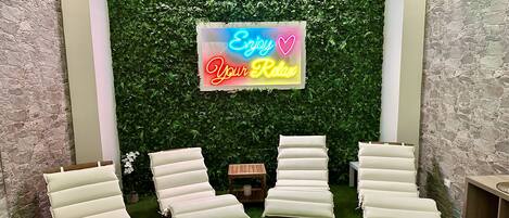 A relaxation area with four white lounge chairs, a neon sign reading "Enjoy Your Relax," and a green wall with foliage.