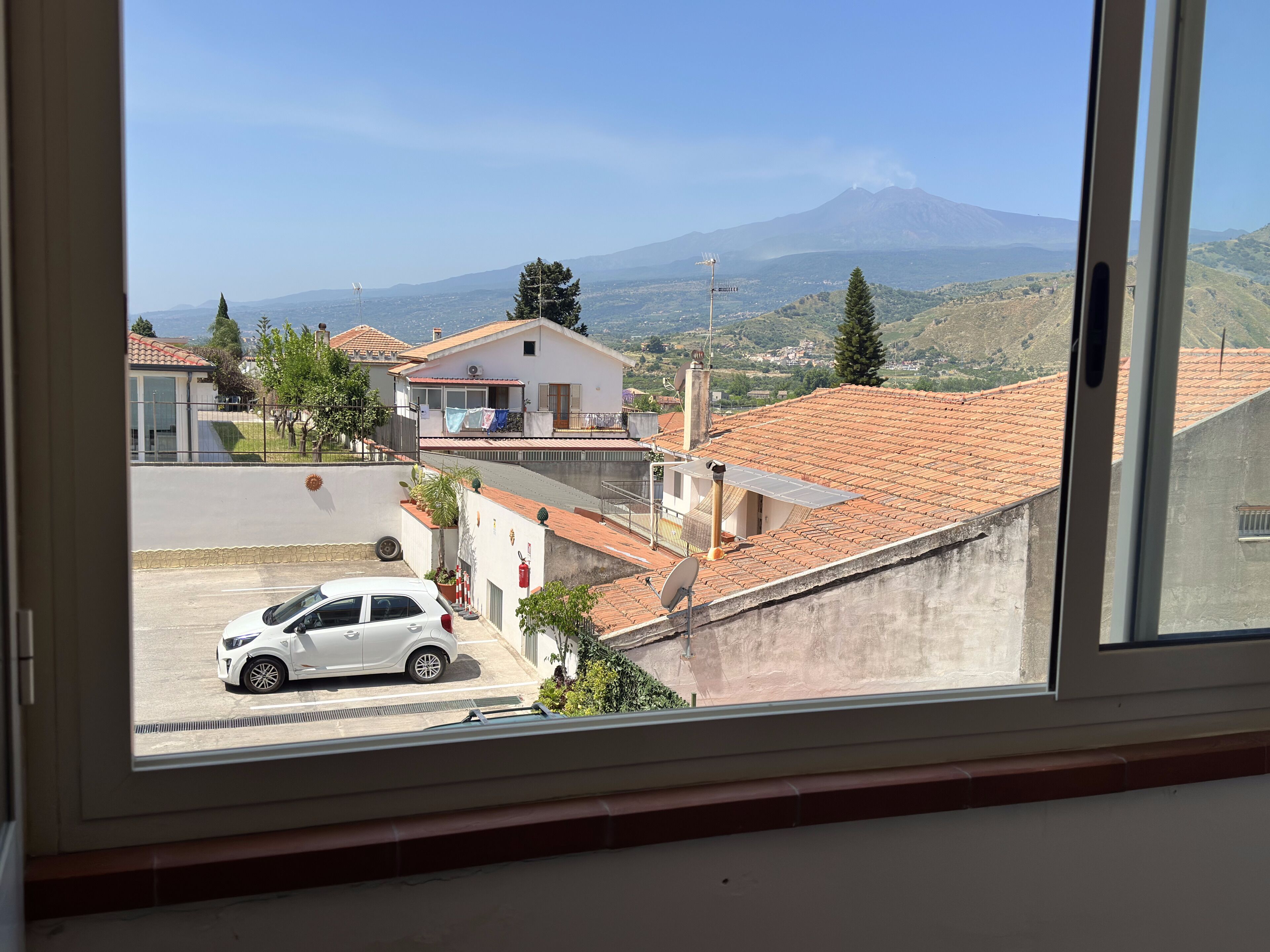A residential area with a view of a mountain, houses with red-tiled roofs, and a white car parked in the foreground.