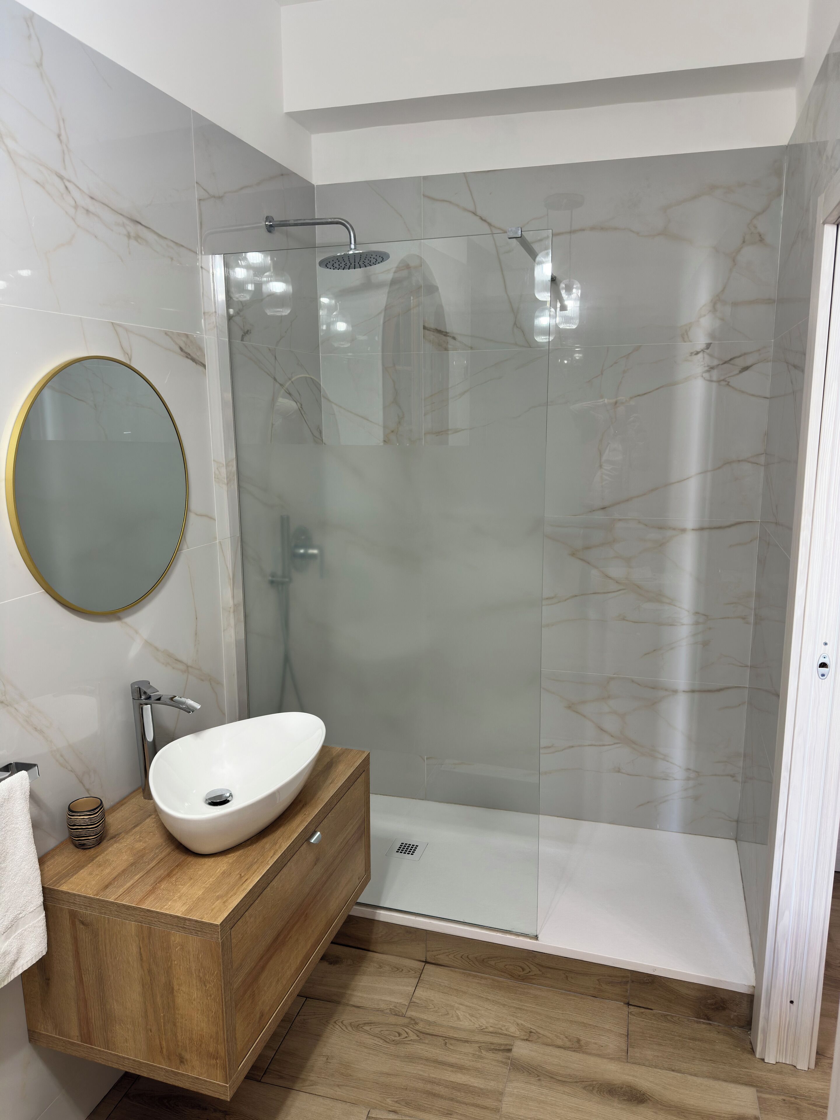 A modern bathroom with a marble-tiled shower enclosure, a wooden vanity with a sink, and a round mirror.