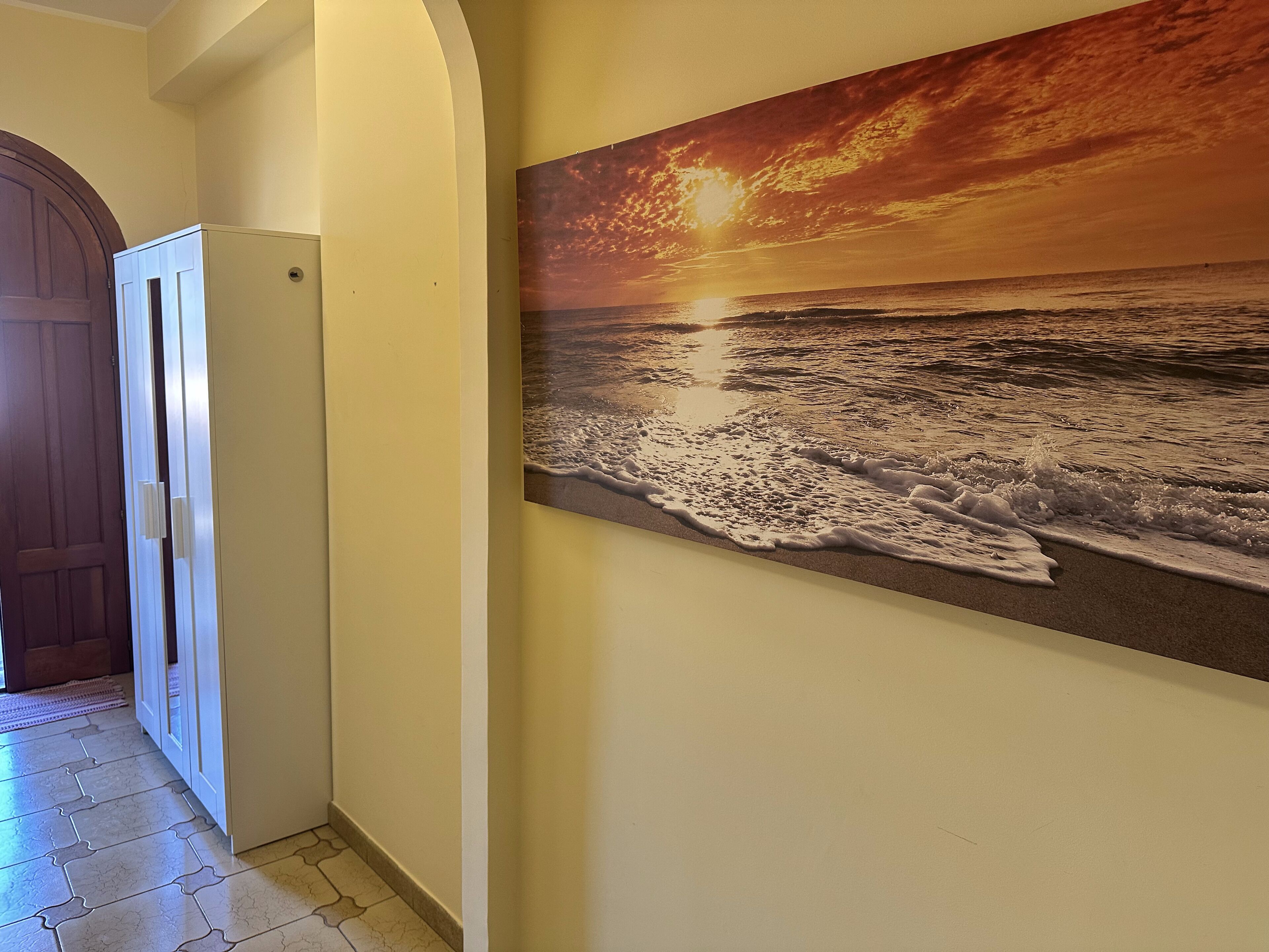 Un couloir avec une porte donnant sur une pièce, un réfrigérateur et un tableau accroché au mur représentant un coucher de soleil sur l’océan.