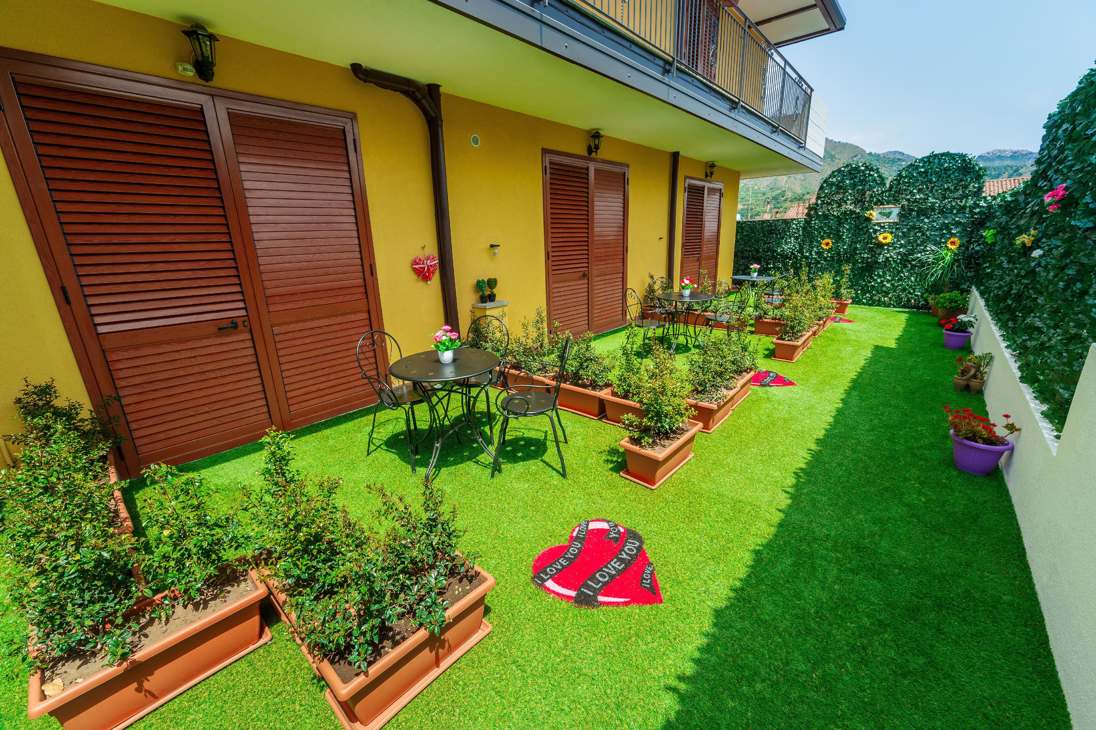 Un patio con césped verde, plantas en macetas, una mesa con sillas y un tapete con forma de corazón que dice 