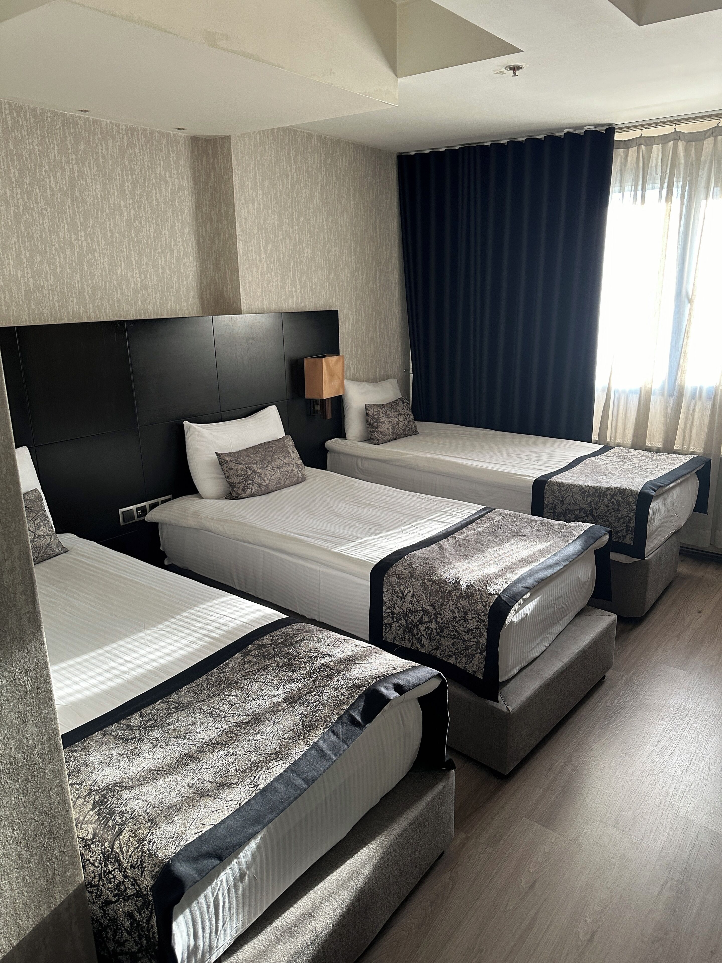 Een hotelkamer met drie op een rij staande bedden, een groot raam met gordijnen en een modern hoofdeinde.
