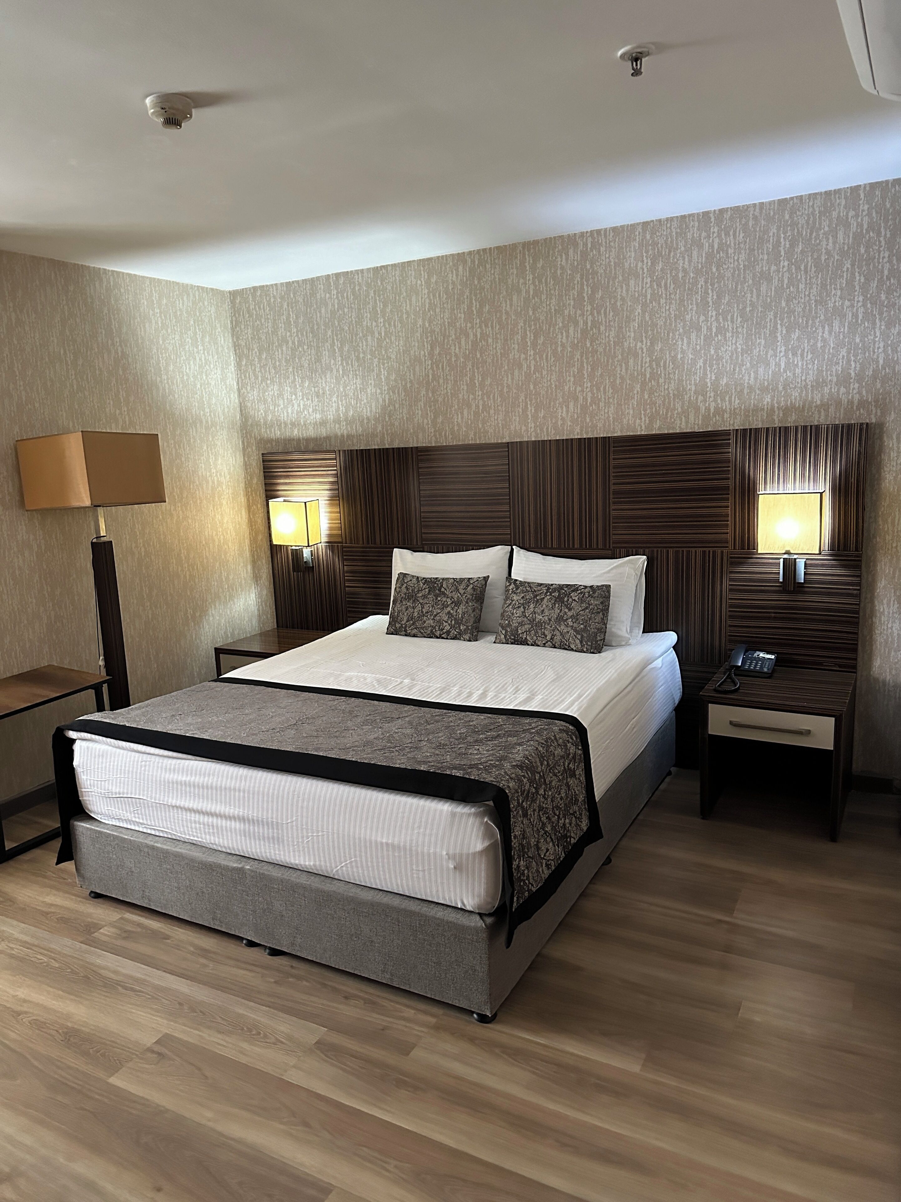 A modern hotel room with a large bed, two bedside tables, a lamp, and a ceiling-mounted smoke detector.