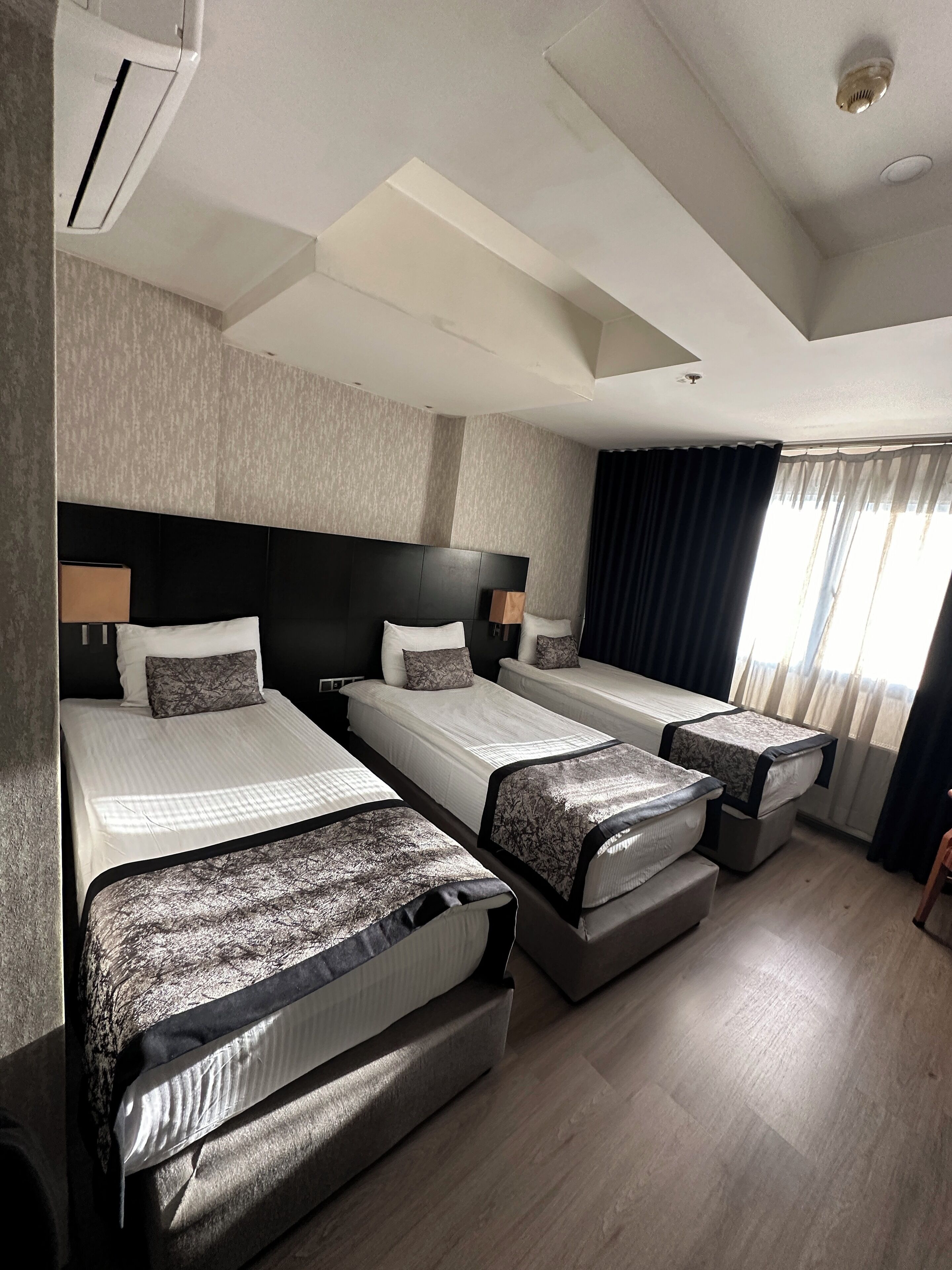 A hotel room with three beds, a headboard, a ceiling light, and a window with curtains.