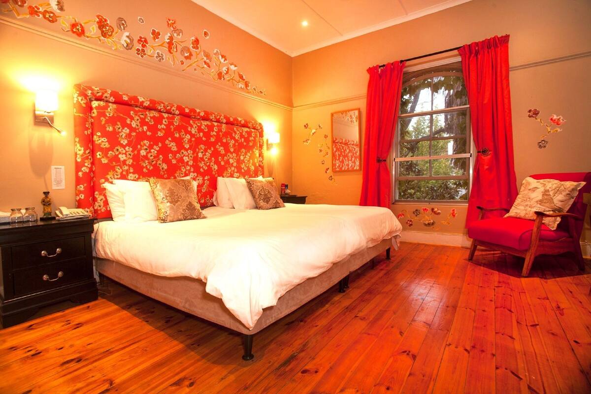 A bedroom with a large bed, a nightstand, a red chair, and a window with red curtains.