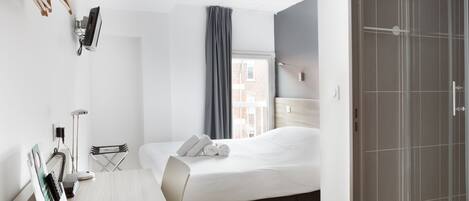 A hotel room with a bed, a desk with a chair, a desk lamp, a telephone, and a window with curtains.