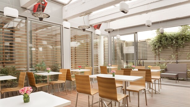 An outdoor dining area with white tables and wooden chairs, large windows, and hanging pendant lights. - Lille City Hotel (Lille)