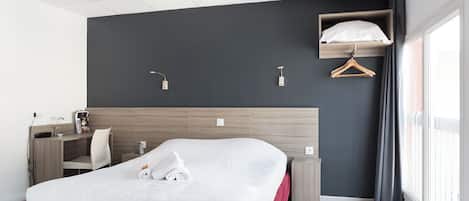A modern hotel room with a bed, bedside table, desk, chair, and a hanging wardrobe.