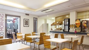 A modern dining area with white tables and wooden chairs, a counter with a coffee machine, and a view of the city through a large window. - Lille City Hotel (Lille)