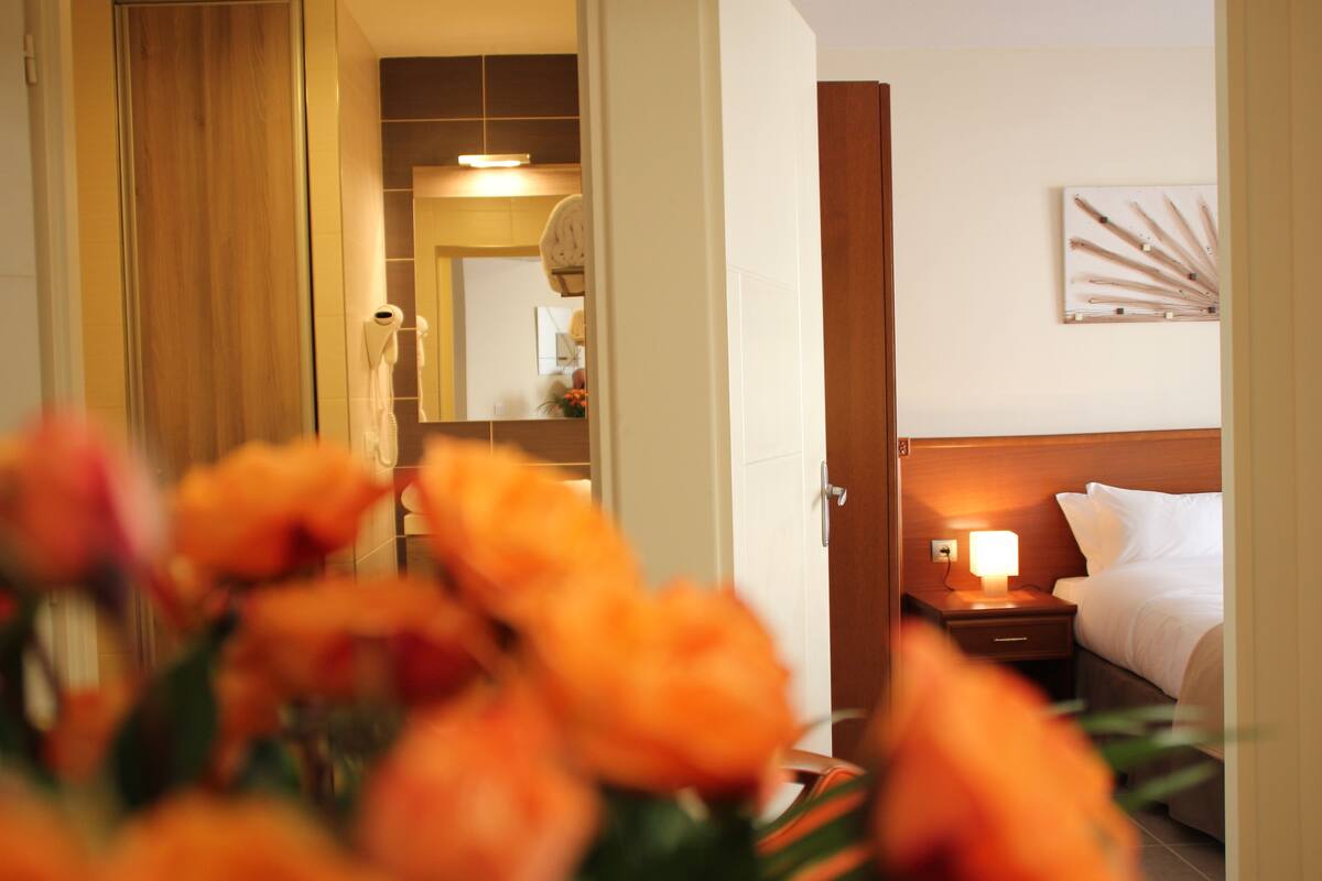 A hotel room with a bed, bedside table with a lamp, a mirror, and a wall art piece.
