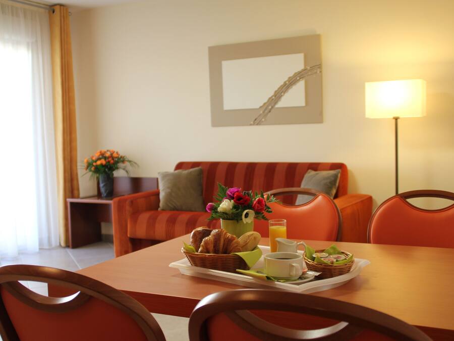 A dining area with a table set for breakfast, including croissants, juice, and coffee. There is a sofa and chairs in the background, and a lamp on the table.