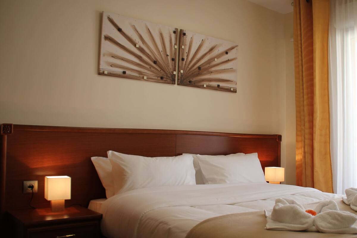 A hotel room with a large bed, two bedside lamps, a wall art piece, and a window with curtains.