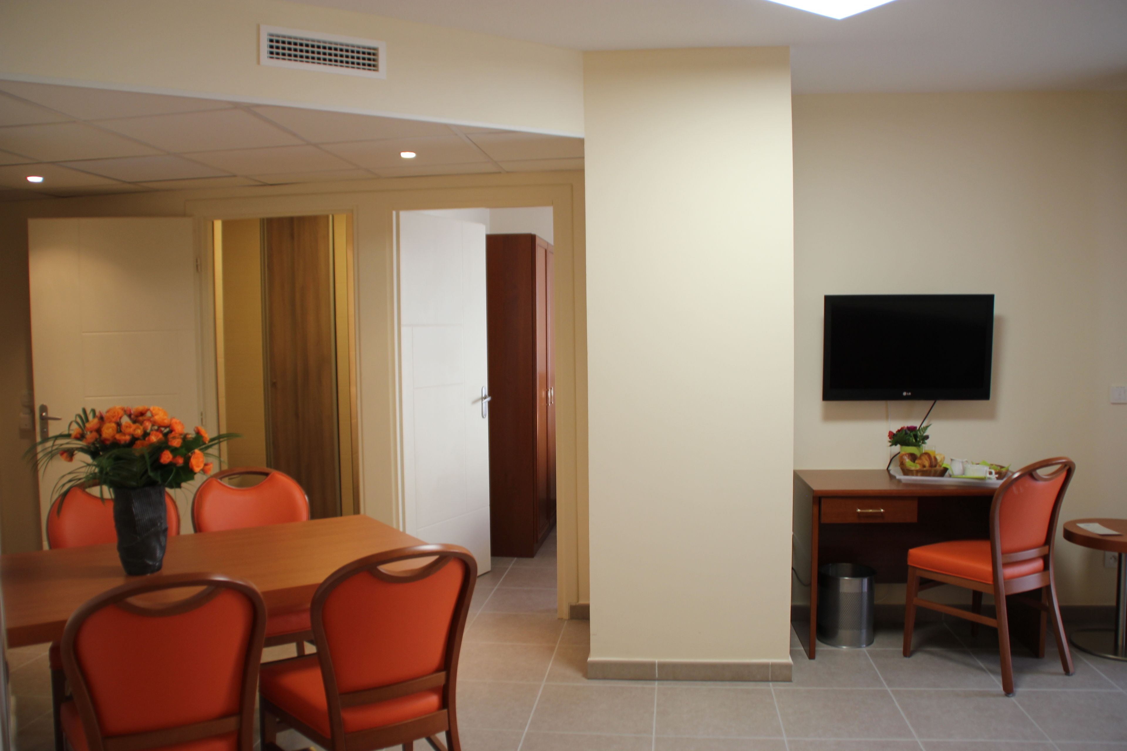 A dining area with a wooden table, orange chairs, and a vase of flowers. There is a TV mounted on the wall and a trash can near the table.