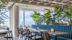 Un restaurante moderno con techos altos, amplias ventanas y vistas a la ciudad. Hay varias mesas y sillas, y las paredes están decoradas con grandes plantas verdes.