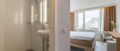 Habitación de hotel con ducha, lavamanos, cama, escritorio y vista a la ciudad.