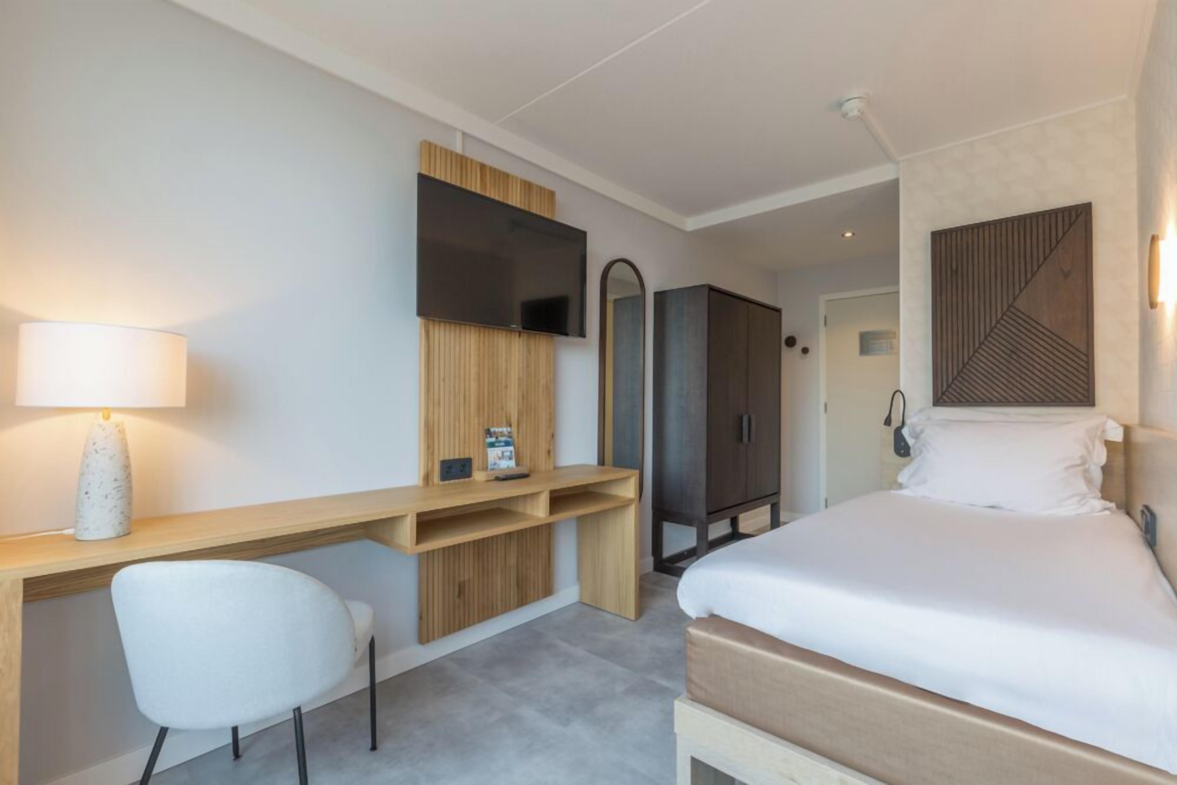 A hotel room with a bed, a desk with a chair, a TV, a wardrobe, and a mirror.