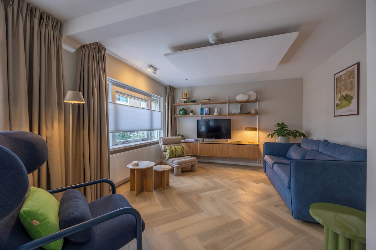 Ein modernes Wohnzimmer mit einer Couch, einem Sessel, einem Hocker, einem Fernseher und einer Küchenzeile mit Regalen und einer Pflanze.