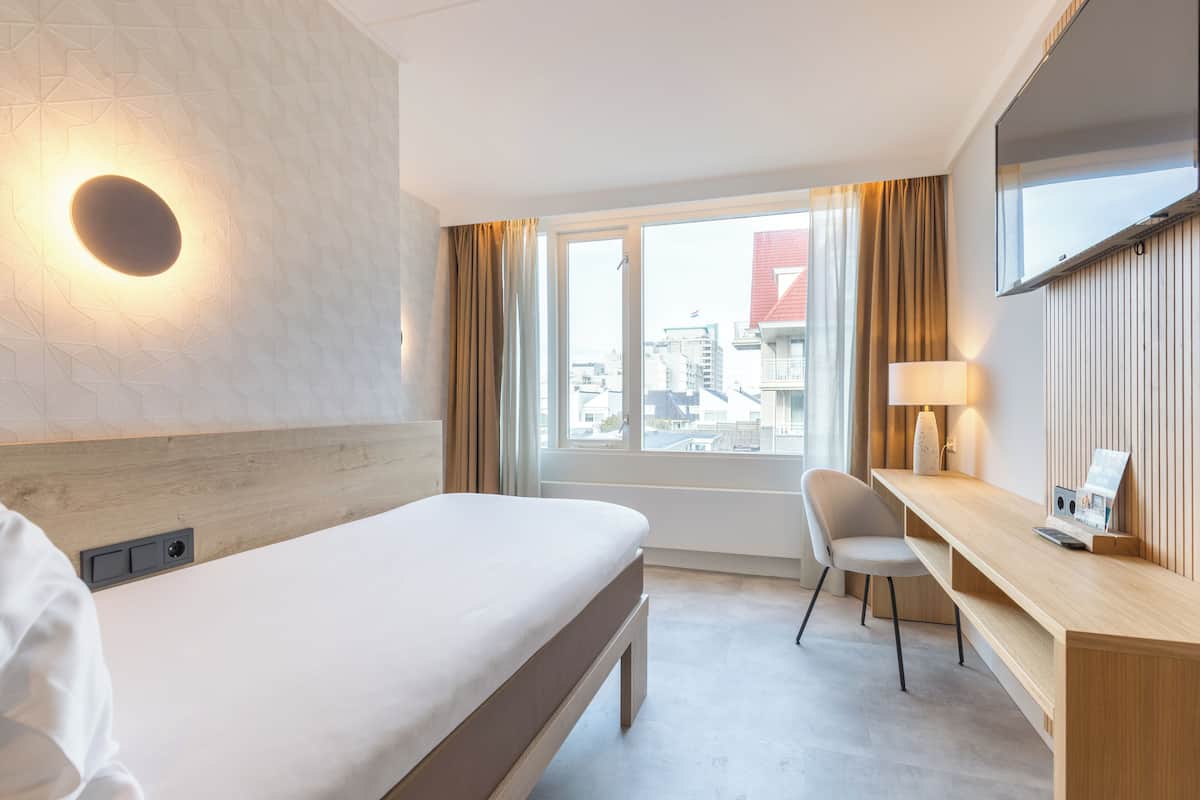 Ein Hotelzimmer mit Bett, Schreibtisch, Sessel, Fernseher und einem fenster mit Vorhängen.