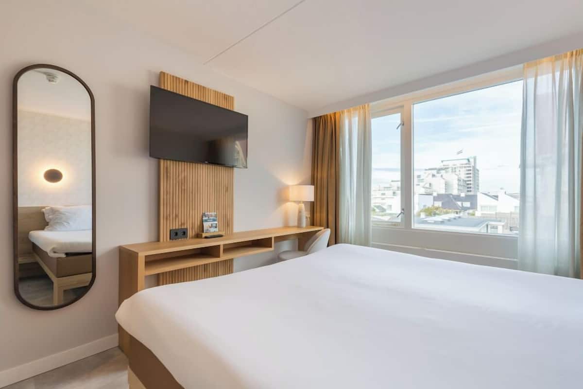 Ein Hotelzimmer mit einem großen Bett, einem an einer Holzvertäfelung montierten Fernseher, einem Nachttisch mit Lampe und Blick auf die Stadt durch große Fenster.