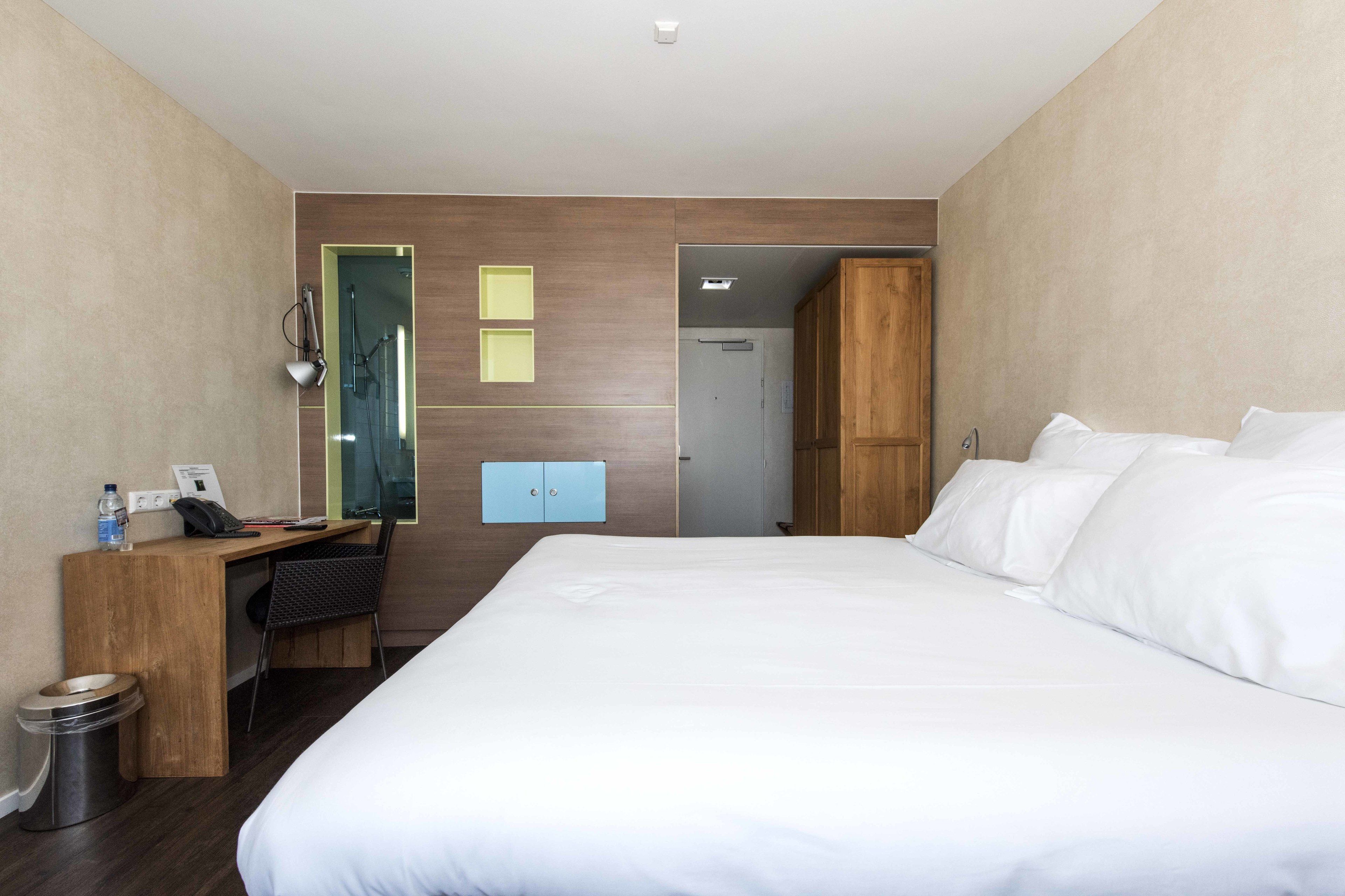 A hotel room with a large bed, a desk with a chair, a small trash bin, and a wardrobe.