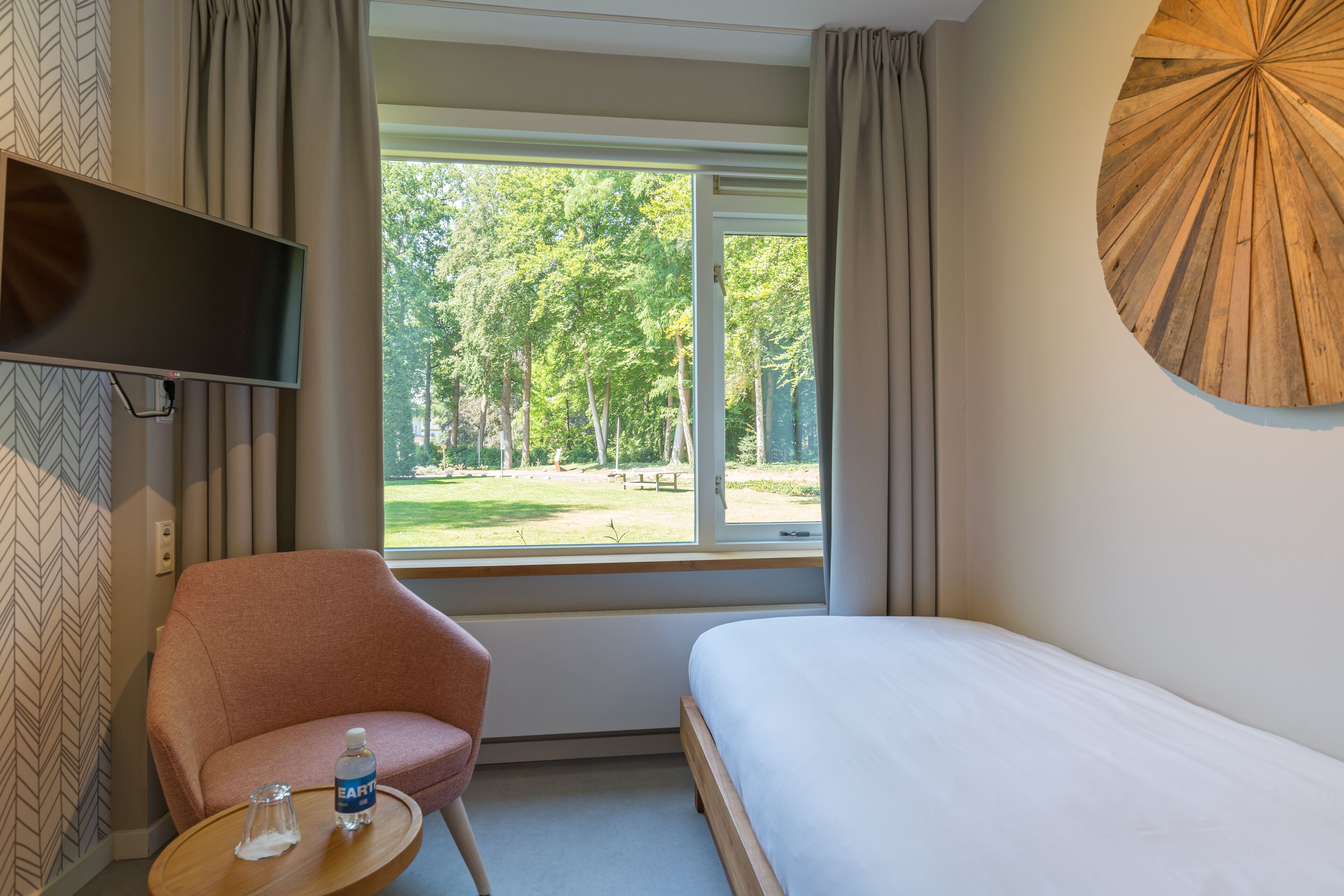 Una camera d'albergo con un letto, una sedia, una televisione, una finestra vista alberi e una decorazione a parete circolare in legno.