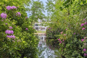 A serene garden with a pond reflecting a white building, surrounded by lush greenery and purple flowers. - Landgoed de Horst (Driebergen)