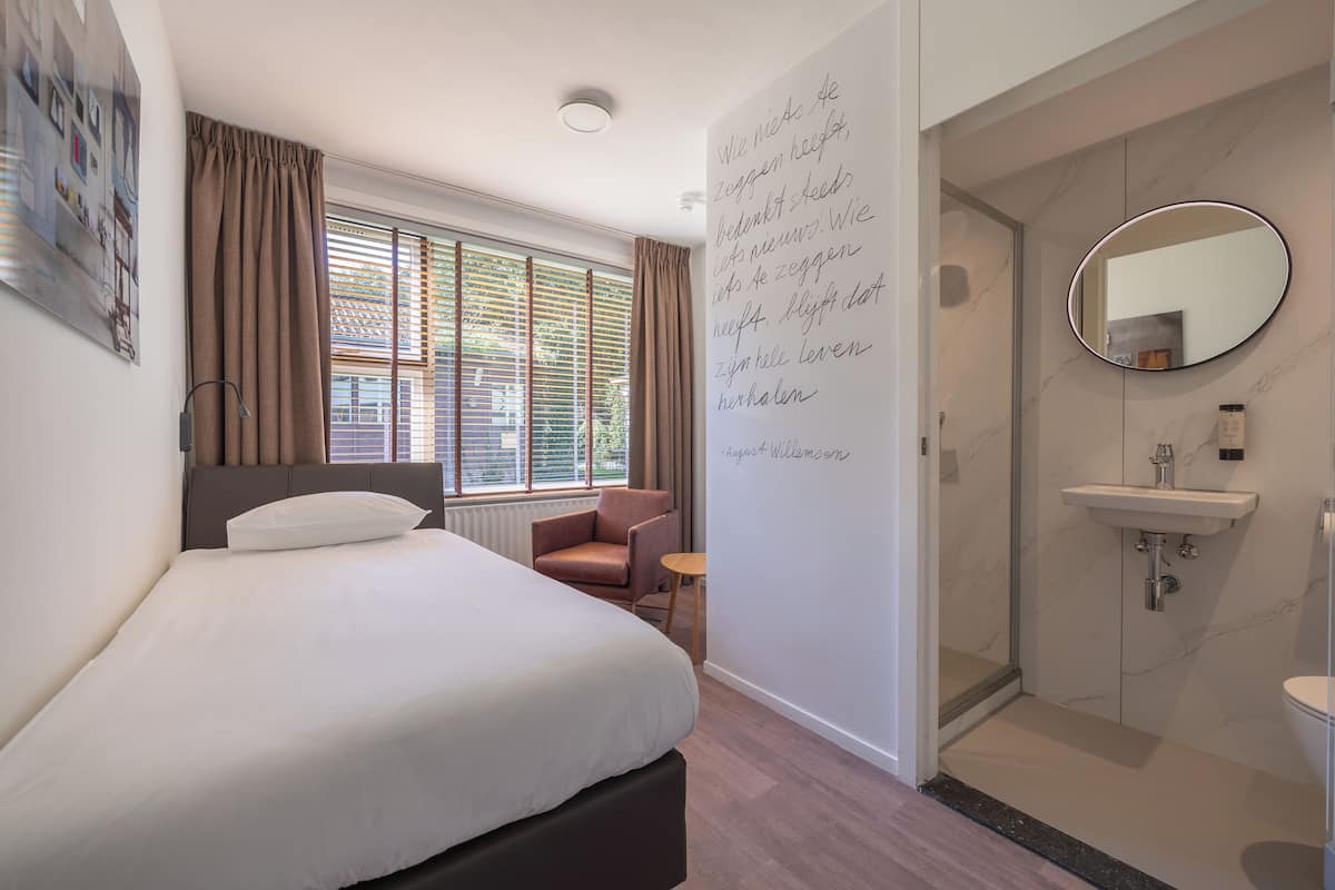 A hotel room with a bed, a chair, a small table, a bathroom with a sink and mirror, and a wall with text.