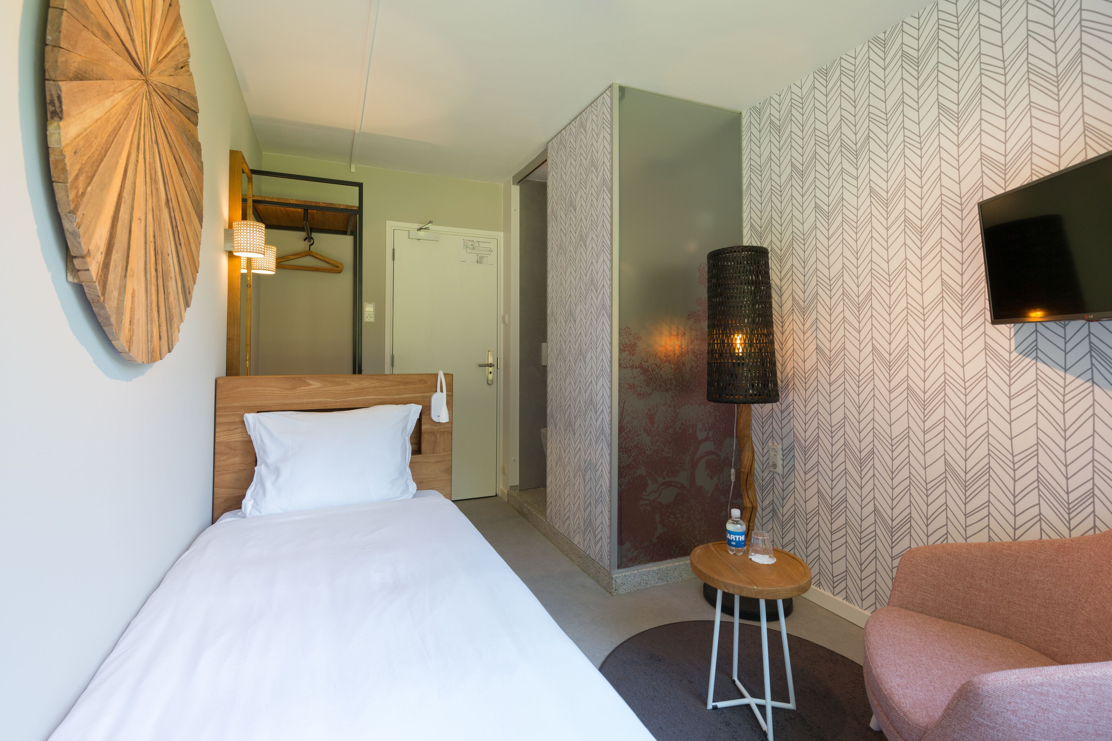 Une chambre d’hôtel avec un lit, une table de chevet avec une lampe, un téléviseur fixé au mur et un papier peint à motifs.