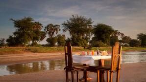 A table set up on a sandy beach with chairs, overlooking a body of water and trees in the background. - Elephant Bedroom Camp (Samburu)