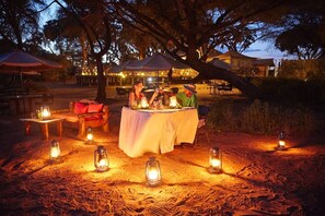 Un espacio de comedor al aire libre con mesas dispuestas bajo un árbol grande, rodeado de faroles.