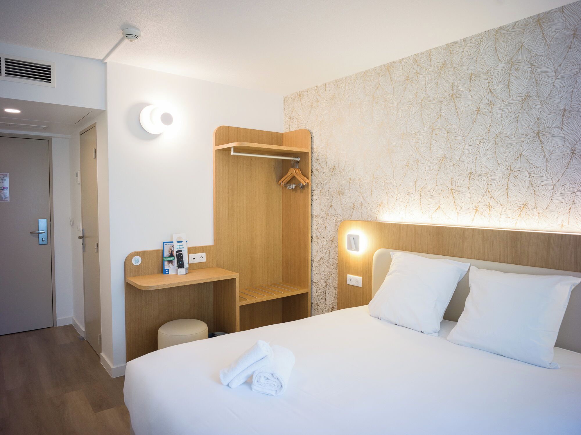 Une chambre d’hôtel comprenant un lit, une table de chevet, une armoire en bois, un espace douche et un luminaire fixé au mur.