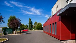 A red building with a 'BIENVENUE | WELCOME' sign, a parking lot, and a red car parked in front. - BRIT HOTEL CONFORT ROISSY SAINT-WITZ (Saint-Witz)