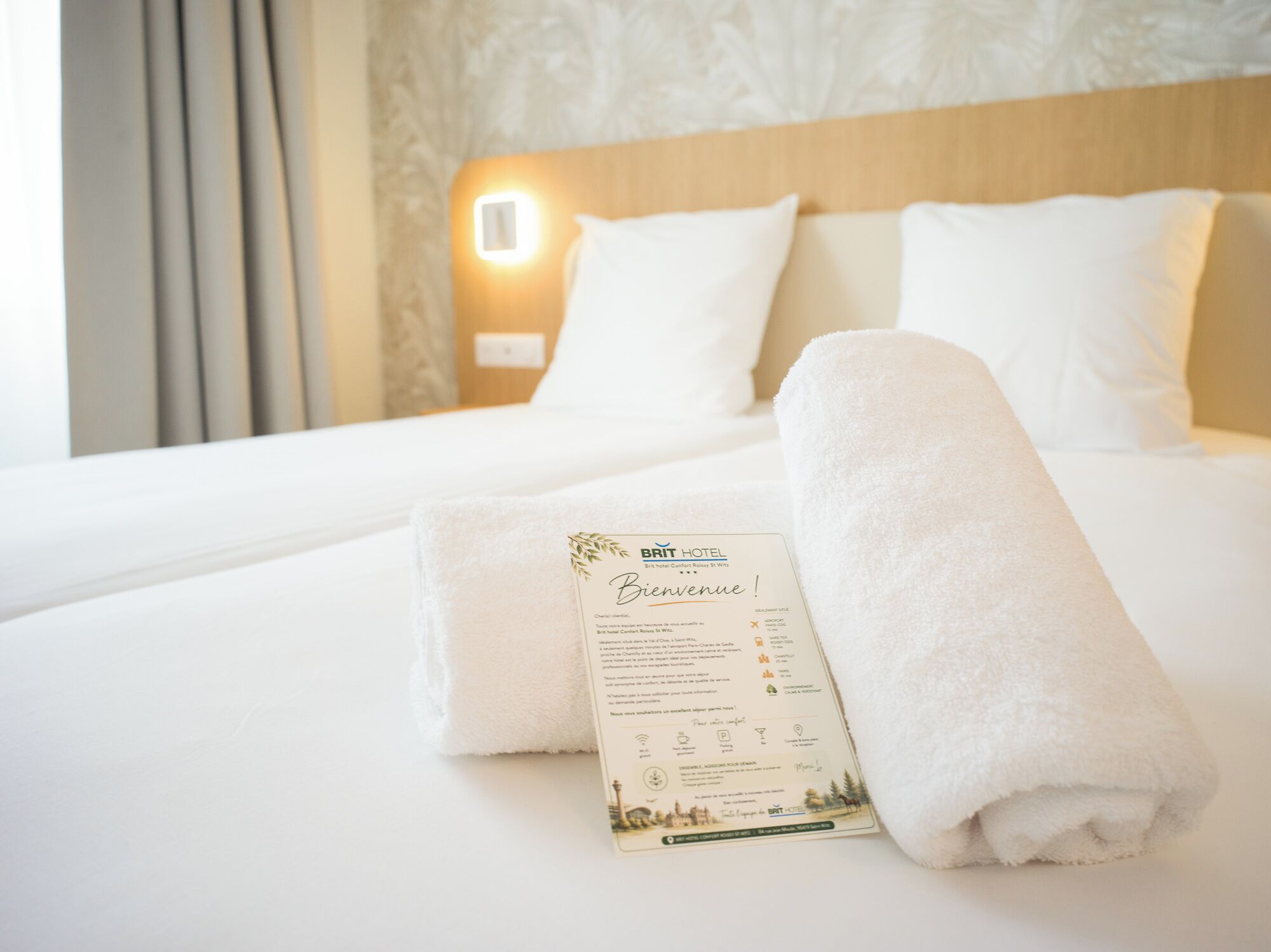 A neatly made bed with white linens, a rolled towel, and a welcome card from the Brit Hotel.